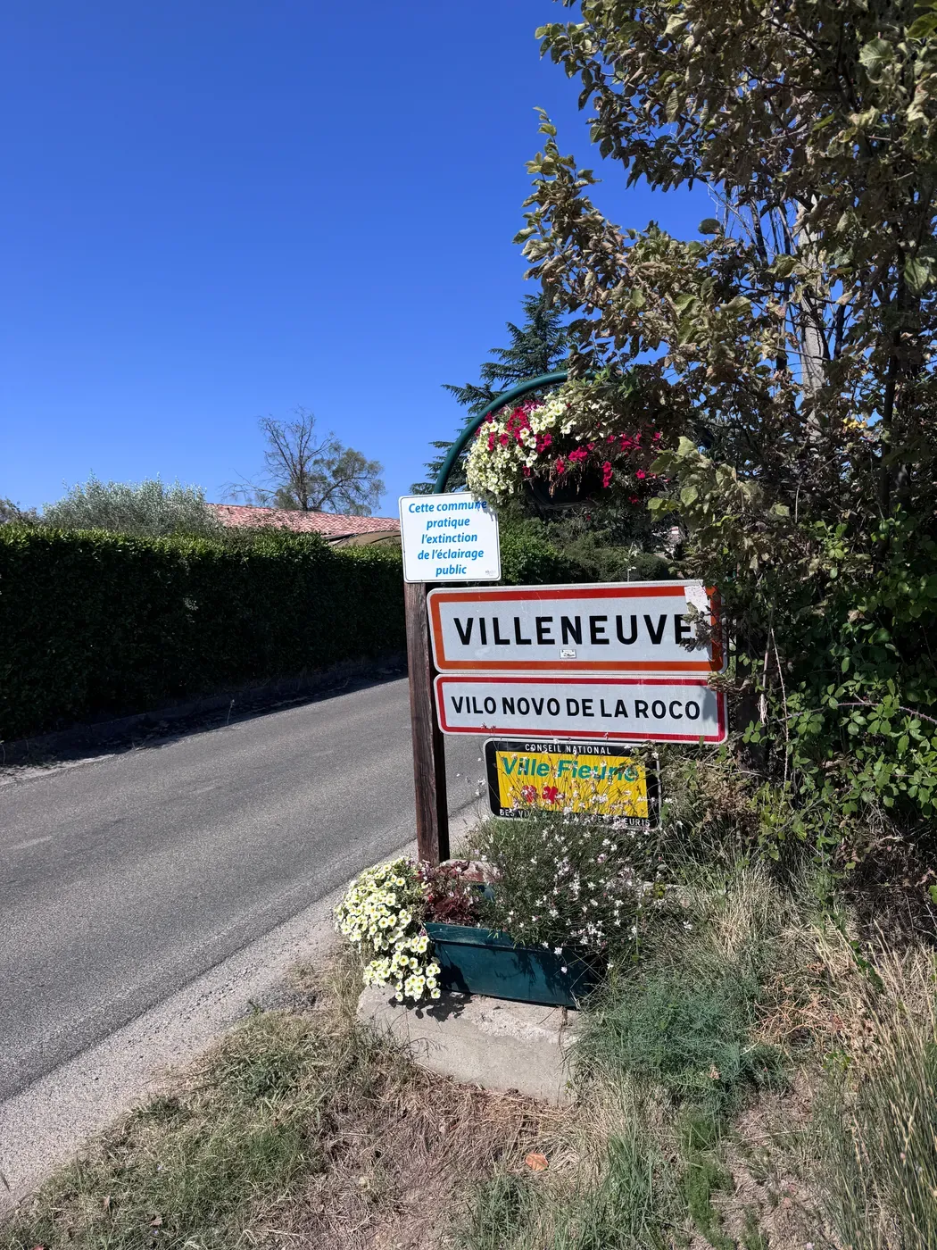 Le panneau d’entrée de Villeneuve, portant aussi le nom occitan Vilo Novo de la Roco, entouré de fleurs.